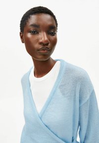 Lichtblauw, doorschijnend gebreid vest met een V-hals en losse mouwen. Draagt over een wit, geribbelde shirt met ronde hals. Gladde textuur.