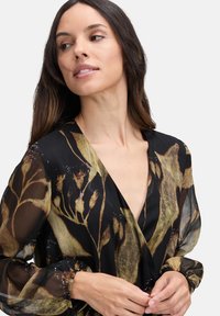Frau mit langen dunklen Haaren, die eine schwarze transparente Bluse mit braunen und goldenen Blumenmustern trägt und leicht nach links schaut.