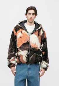 Giacca con cappuccio pelosa in marrone, arancione e crema con chiusura a zip, tessuto texturizzato e vestibilità rilassata, abbinata a un maglione bianco e jeans blu.