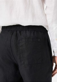 Zwarte linnen broek met een elastische tailleband, voorzien van een enkele achterzak en een zachte, gestructureerde stof. Gdraag met een wit shirt.