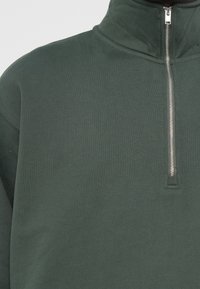 Mørkegrøn halvzip pullover lavet af ribbet stof. Har en sølvfarvet lynlås og en høj krave for ekstra varme.