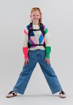 Young girl with red hair wearing a colorful striped sweater, blue jeans, pink sneakers, and a black backpack with chest and waist straps.