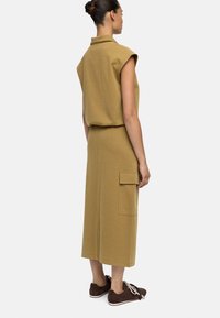 Szydełkowy, musztardowy zestaw składający się z bezrękawnika i spódnicy do połowy łydki z boczną kieszenią, zestawiony z brązowymi trampkami.