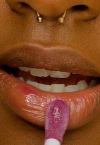 Primo piano di labbra con un finish lucido, che mostrano una tonalità marrone chiaro. Un applicatore soffice con una sfumatura rosa viene utilizzato sul labbro inferiore.