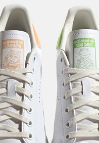 Bílé kožené tenisky Adidas Stan Smith mají oranžové a zelené patní záložky s postavičkami na jazyku. Kulaté tkaničky, texturovaný povrch.