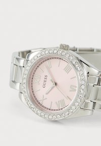 Ceas de mână Guess argintiu cu cadran roz, lunetă cu cristale, cifre romane și brățară metalică cu zale, indicând ora 10:53:39.
