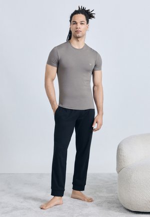 Moški z dredami stoji bosi na preprogi, oblečen v oprijeto sivo majico in črne športne hlače, ob njem je ukrivljeno kremno naslanjač.