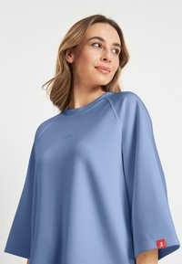 Lichtblauwe oversized sweatshirt met een gladde textuur, voorzien van driekwartmouwen en een rode accenttag op de linker mouw.
