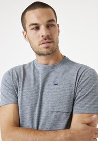 Grau gestreiftes Kurzarm-T-Shirt mit Rundhalsausschnitt, das eine Brusttasche und ein kleines Markenlabel auf der Tasche aufweist. Weicher Stoff.