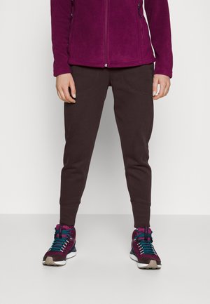 Person iført mørkebrune joggingbukser, magenta lynlåsjakke og matchende magenta og blågrønne vandresko står på en neutral baggrund.