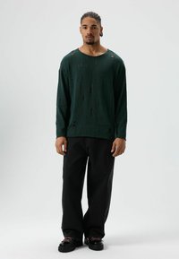 Groene trui met lange mouwen en een losse pasvorm, met meerdere beschadigingen. Gecombineerd met zwarte losse broek en grove zwarte schoenen.