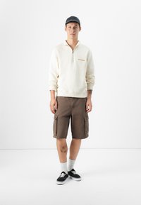 Creme halv-zip sweatshirt med broderet detalje, brune cargo shorts med sidelommer, hvide crew sokker og sorte sneakers.
