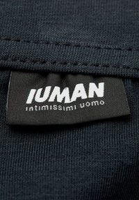 Zwarte stoffen label met witte letters waarin "IUMAN" en "intimissimi uomo" staat. Het label is genaaid op een gestructureerde donkere stof.