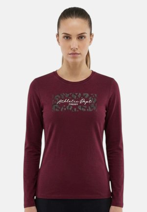 Donna che indossa una maglia a maniche lunghe color granata con "Athletic Dept Freddy" e una stampa leopardata sul petto, in piedi davanti a uno sfondo semplice.