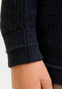 Dunkelblauer gestrickter Pulloverbund mit rippenförmiger Struktur und gerader Kante, wobei eine Hand gegen einen neutralen Hintergrund ruht.