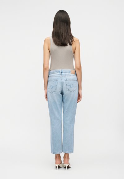 Γυναίκα με ίσια σκούρα μαλλιά φορώντας μπεζ αμάνικο τοπ, ανοιχτό μπλε ψηλόμεσο τζιν και λευκά παπούτσια με τακούνι, όρθια με την πλάτη προς το μέρος μας.