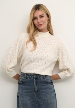 Pull en tricot couleur crème avec un col montant et des manches bouffantes, présentant un motif texturé. Associé à une jupe en denim.