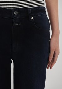 Nahaufnahme von dunkelblauen Jeans mit Knopf- und Taschendetails, getragen mit einem schwarz-weiß gestreiften Oberteil, Hand seitlich am Körper.