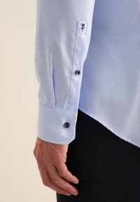 Camicia a maniche lunghe azzurro chiaro con una texture liscia, dotata di bottoni neri e un design ricamato sul polsino, forma del polsino esagonale.