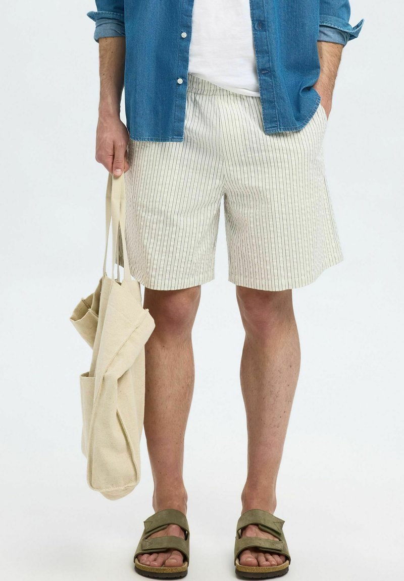 Shorts rayés pour hommes en beige clair et crème, fabriqués en tissu léger. Assortis à une chemise bleue et des sandales, tenant un sac fourre-tout en toile.