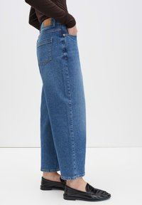 Wide-leg blue denim jeans with a textured finish, high waist, and back pockets. Paired with black loafers for a casual look.