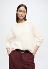 Krémszínű pulóver kerek nyakkal és laza szabással, bordázott mandzsettákkal, bordó nadrág fölött viselve, fekete övvel arany csattal.