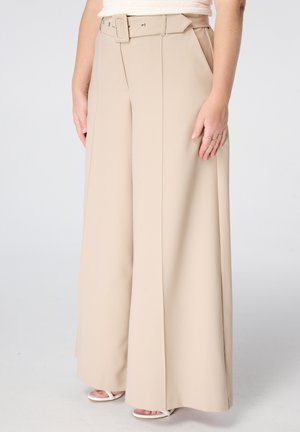 Femme portant un pantalon beige taille haute à jambes larges avec ceinture assortie et des sandales blanches à bouts ouverts, debout devant un fond uni.