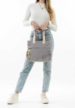 Femme en pull blanc et jean bleu clair tenant un sac fourre-tout gris avec des poignées beiges et un tire-zip jaune.