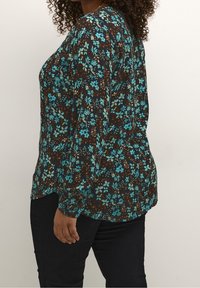 Blouse fleurie à manches longues, avec un fond marron foncé et des fleurs turquoise. Texture lisse, ourlet arrondi et coupe décontractée.