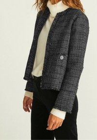Schwarze und weiße Tweed-Shortjacke mit Rundhalsausschnitt, fransigen Kanten, einem Knopfverschluss und einer Detailtasche auf der Vorderseite.