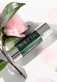 Bouteille verte avec un couvercle argenté, étiquetée "Oskia London City Life Concentrate." Entourée de pétales de fleurs roses et de feuilles vertes.