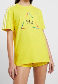 Gul bomullströja med korta ärmar, med en triangulär grafik som har "Hu" i svart och regnbågsfärger som accent. Vita ränder på axlarna.