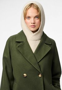 Abrigo de lana verde con doble botonadura dorada, solapas amplias, acompañado de una bufanda de punto color crema que envuelve el cuello y la cabeza.