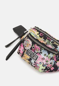 Sac zippé à motif floral noir avec des détails en œillets argentés, arborant un logo "Versace Jeans Couture" et une bandoulière amovible pour le poignet.