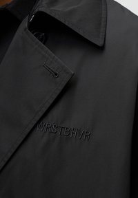 Abrigo negro con tejido texturizado, gran collar y logo bordado "WRSTBHVR" en el lado izquierdo. Cierre de botón estándar visible.