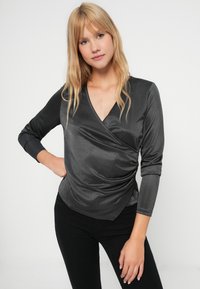 Šedý zavinovací top vyrobený z lesklého materiálu, má výstřih do V, dlouhé rukávy a nařasené detaily v oblasti pasu, je kombinován s černými kalhotami.