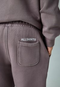 Bikšu sporta bikses izmutētas mauva krāsas audumā ar teksturētu apdari, ar aizmugures kabatu un baltu "ALLSAINTS" logotipu. Redzama elastīga jostas detalizācija.