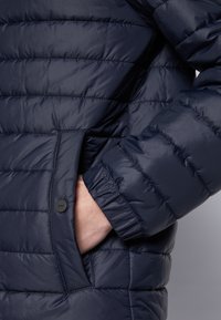 Marineblå pufferjakke med vandret quiltet mønster, letvægtsmateriale, elastiske manchetter og en knaplukket sidelomme med et mærkelogo.