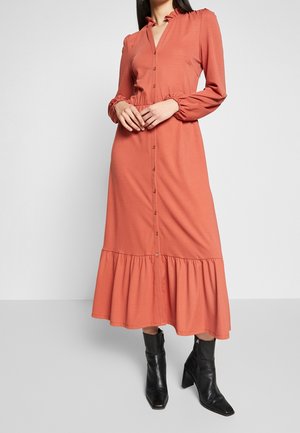 Kvinde iført en langærmet, knappet midi-kjole i rust-orange med flæsekant og sorte ankelstøvler med hæl.