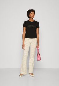 Camiseta negra ajustada con logo en relieve "GUESS", pantalones de campana crema con estampado y zapatos blancos tipo slip-on. Bolso de mano de cuadros rosas con correa corta.