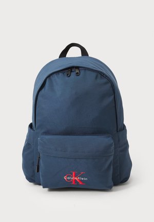 Sac à dos bleu marine fabriqué en tissu durable, avec deux compartiments zippés, une poche avant et un accent logo Calvin Klein rouge.