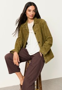 Woman with long hair wearing an olive green suede jacket, white lace top, and brown pants, seated on a wooden stool against a plain background.