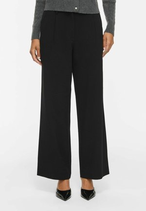 Femme portant un pantalon noir taille haute à jambes larges, associé à des chaussures noires à bouts pointus et un pull gris boutonné.