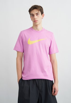 Joven con pendientes que lleva una camiseta rosa con un gran logotipo amarillo de Nike y pantalones negros holgados, de pie frente a un fondo liso.
