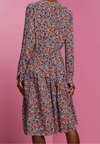 Nainen, joka on pukeutunut pitkähihaiseen, polvimittaisiin mekkoihin, joissa on purppuran, punaisen, mustan ja valkoisen kukkakuosiset kankaat vaaleanpunaisella taustalla.