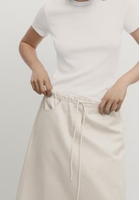 Hvid tætsiddende t-shirt parret med en lys beige nederdel med elastisk talje, snørebånd og sidelommer. Glat tekstur, enkelt design.