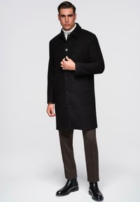 Schwarzer Wollmantel mit Kragen, Knopfverschluss und Seitentaschen. Getragen über einem cremefarbenen Rollkragenpullover und kombiniert mit dunklen Hosen und Stiefeletten.