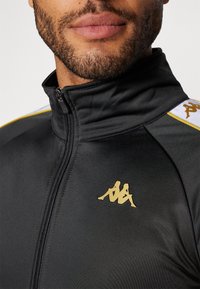 Melna sporta jaka ar rāvējslēdzēju un faktūru. Tiek piedāvāts ar zeltītu Kappa logotipu un dzeltenām akcenta svītrām uz pleciem.
