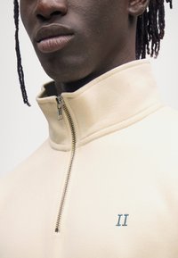 Pull-over beige à demi-fermeture éclair en tissu doux ; doté d'une fermeture éclair en métal et d'un petit "II" bleu brodé sur la poitrine.