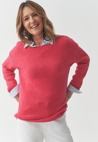 Maglione rosa lavorato a maglia con una vestibilità ampia, scollatura larga e maniche lunghe. Sotto si trova una camicia azzurra con colletto. Indossato con pantaloni bianchi.